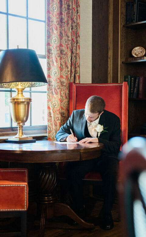a groom writing his wedding vows in a luxury hotel room study with dark wood bookshelf and table