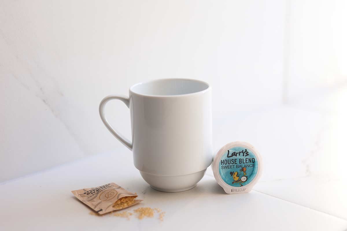 white coffee mug next to an open single sering packet of brown sugar and a house blend coffee pod
