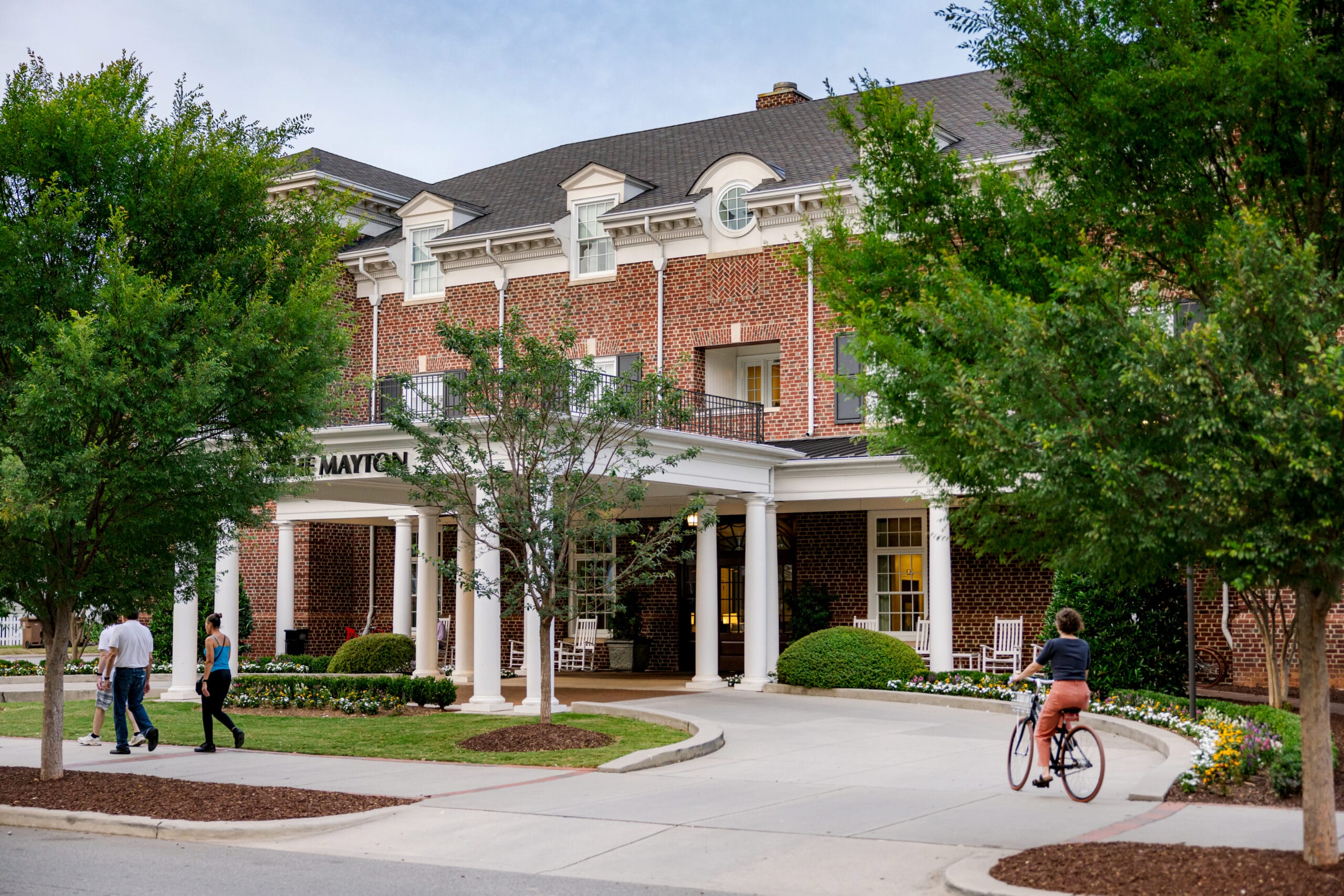 Exterior view of The Mayton brick hotel building featuring white columns at the entrance, lush green trees, pedestrians walking on the sidewalk, and a person riding a bicycle along the driveway.