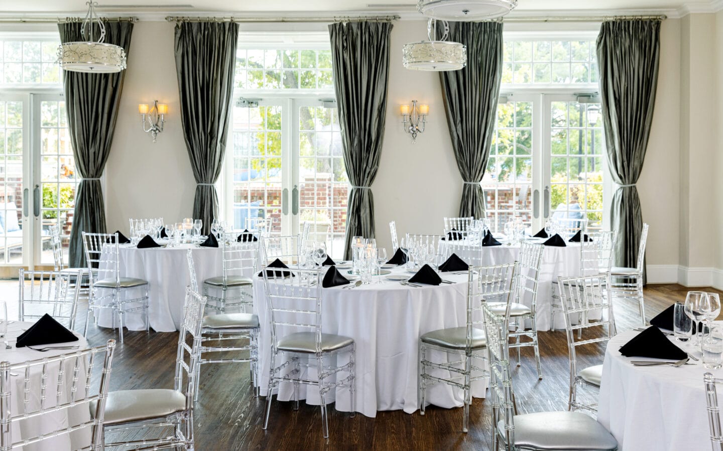 An elegant indoor reception hall set with round tables, white tablecloths, clear Chiavari chairs, dark napkins, and wall-mounted chandeliers in front of large windows framed by dark drapes.