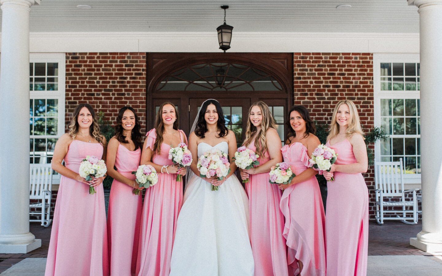 A bride in a white strapless wedding gown holding a floral bouquet, standing in front of a brick building entrance surrounded by six bridesmaids in matching pink dresses.