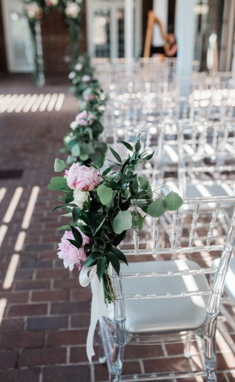 Clear acrylic Chiavari wedding aisle chair decorated with pink roses, white flowers, eucalyptus greenery, and a white ribbon bow along a brick walkway.