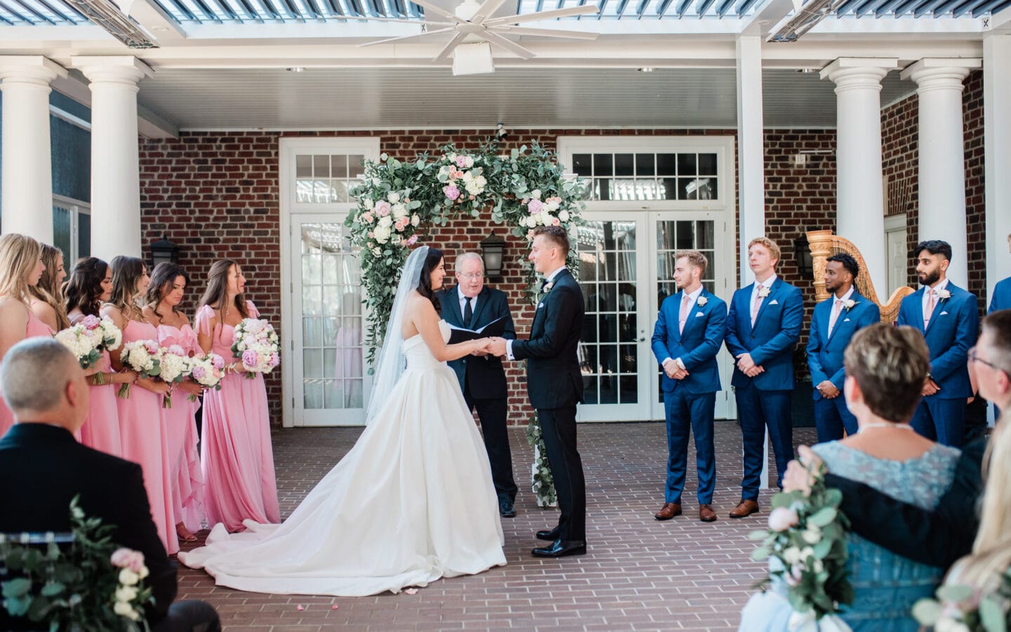 A bride and groom holding hands at the altar during an outdoor wedding ceremony, flanked by bridesmaids in pink dresses, groomsmen in blue suits, and an officiant under a floral archway.