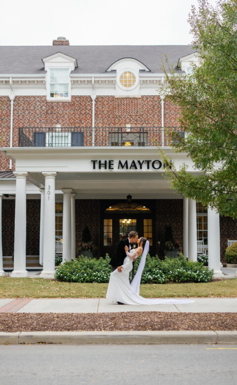 A bride and groom kissing outdoors in front of The Mayton hotel, with white pillars and a brick facade in the background.