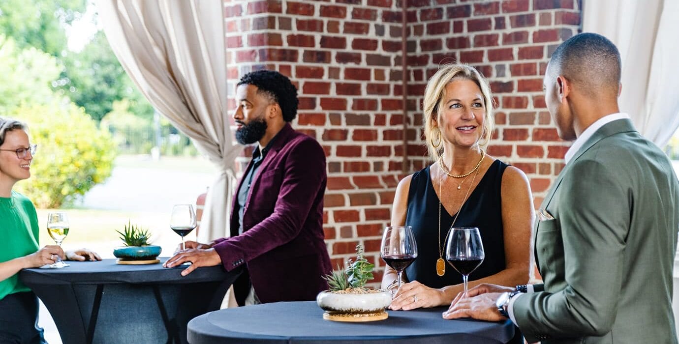 Four elegantly dressed guests socializing around tall cocktail tables with wine glasses on an outdoor brick patio with white curtains.